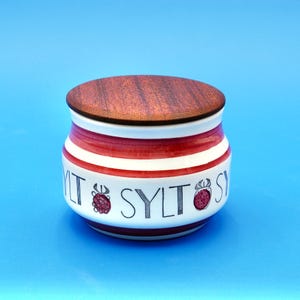 Op de afbeelding: Een kleine, ronde keramische pot met een houten deksel. De pot is wit met rode strepen en het woord "SYLT" in zwarte letters. De pot heeft een bessenontwerp en staat tegen een blauwe achtergrond.