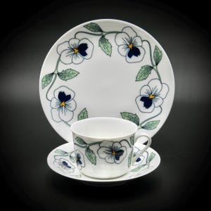 Puede incluir: Un juego de taza y platillo de té blanco con un diseño floral. La taza y el platillo tienen un borde de enredaderas verdes con pensamientos blancos y centros azules.