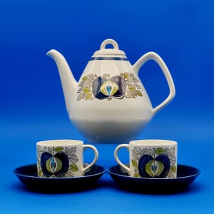 May include: A white teapot with a blue and green apple design and two matching teacups and saucers. The saucers are a deep blue color.