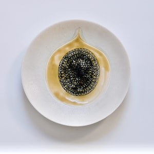 Puede incluir: Un plato de cerámica blanco con un diseño de higo. El higo está en el centro del plato, con un interior amarillo-marrón y un patrón de semillas negro. El plato tiene una pequeña extensión puntiaguda en la parte superior.