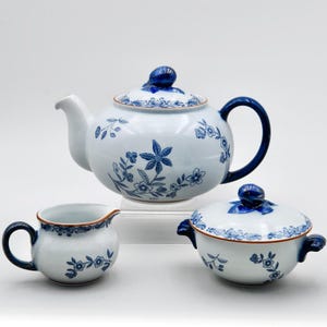 May include: A white porcelain tea set with blue floral designs. The set includes a teapot with a curved handle, a creamer, and a covered sugar bowl. The rims and handles are accented with blue. The teapot lid has a blue finial.