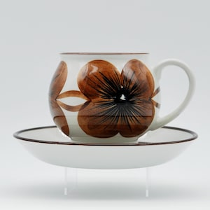 May include: A white ceramic teacup and saucer with a brown floral design. The cup has a rounded shape and a handle. The saucer has a brown rim.