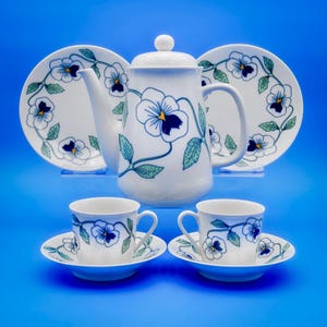 Puede incluir: Un juego de té de cerámica blanca con un diseño floral. El juego incluye una tetera, dos platos y dos tazas con platillos. El patrón floral presenta pensamientos azules y hojas verdes. El fondo es azul liso.