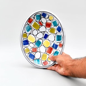May include: Oval ceramic plate with a white base and a colourful, abstract design. The plate features a black rim and is decorated with various shapes in red, yellow, blue, and green. The plate is held by a hand against a white background.