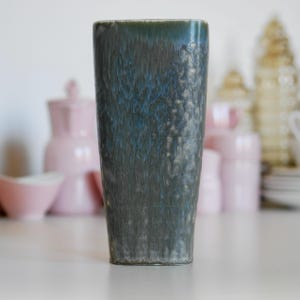 May include: A tall, dark green ceramic vase with a mottled, textured surface. The vase has a rectangular shape and a slightly flared opening.