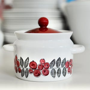 Op de afbeelding: Witte keramische pot met een rood deksel en een zwart en rood cranberry ontwerp. De pot heeft twee handvatten.