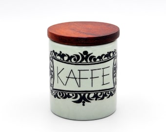 以下が含まれることがあります： 黒と白の装飾的なデザインと黒い文字で「Kaffee」と書かれた白いセラミック製のコーヒー缶。缶には木製の蓋が付いています。