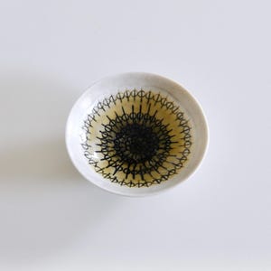 Puede incluir: Un cuenco pequeño y redondo de cerámica blanca con un diseño decorativo negro y amarillo. El interior del cuenco presenta un diseño concéntrico de líneas negras que irradian desde el centro, con un fondo amarillo. El borde es de color beige claro.