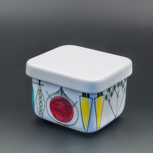 May include: A white ceramic square container with a matching lid. The container is decorated with colorful, hand-drawn illustrations of vegetables, including a red onion, yellow carrots, and green herbs. The design is whimsical and modern.