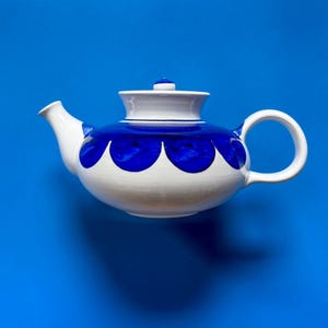 Può includere: Una teiera in ceramica bianca con un corpo largo e arrotondato e un coperchio abbinato. Il design presenta una fascia prominente di grandi motivi semicircolari blu scuro che circondano la sezione centrale, completata da un manico e un beccuccio.