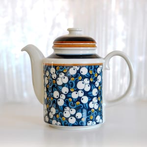 Könnte beinhalten: Eine weiße Keramik-Teekanne mit Deckel. Die Teekanne hat einen blauen Hintergrund mit einem Muster aus weißen Beeren und blauen Blättern. Der Deckel hat ein braunes und schwarzes Band. Griff und Ausguss sind weiß.
