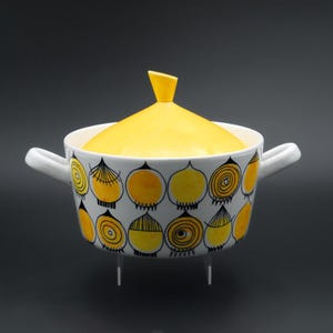 Op de afbeelding: Een witte keramische kom met een gele deksel en handvatten. De kom is versierd met gele en zwarte abstracte ontwerpen. De kom heeft drie kleine witte poten. De deksel heeft een kleine, schuine handgreep.