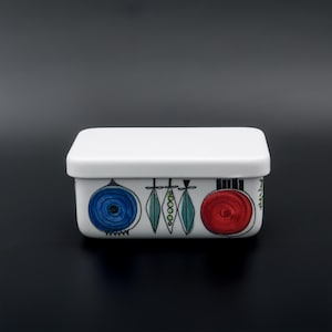 Puede incluir: Una mantequera rectangular blanca de cerámica con tapa a juego. El plato está decorado con ilustraciones coloridas y estilizadas de verduras, incluyendo una cebolla azul, vainas verdes y un tomate rojo. El diseño recuerda a la vajilla de cocina vintage.