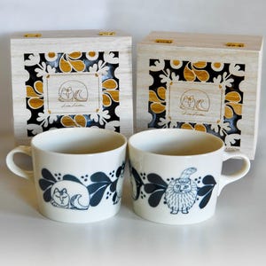 Puede incluir: Dos tazas de cerámica blanca con ilustraciones de gatos azules y un borde decorativo. Las tazas se combinan con dos cajas de madera con un diseño de gato similar y detalles dorados. Las cajas tienen un acabado de madera clara.