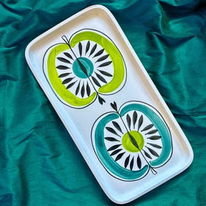Könnte beinhalten: Ein rechteckiges weißes Serviertablett mit einem Retro-Apfel-Design. Das Tablett zeigt zwei stilisierte Apfelhälften in Grün und Türkis, mit schwarzen Akzenten und einem türkisfarbenen Zentrum. Das Tablett ist etwa 35 cm lang und 20 cm breit.