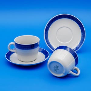 Puede incluir: Tazas y platillos de cerámica blanca con diseño de rayas azules. Las tazas tienen asa y se combinan con platillos a juego. El juego se muestra sobre un fondo azul. Diseño clásico.