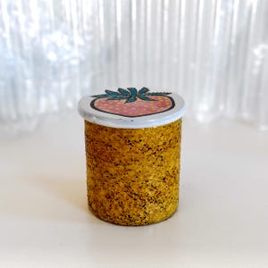 May include: A small, cylindrical jar with a textured, golden-yellow exterior. The jar has a white lid decorated with a strawberry design. The strawberry is pink with green leaves and a black outline. The jar is on a white surface.