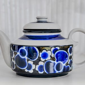 May include: A ceramic teapot with a white body, a gray lid, and a curved handle. The teapot features a blue and white floral pattern on a black background. The lid has a small knob and a black band. The spout is curved.