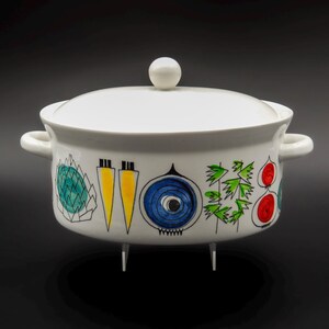 Op de afbeelding: Witte keramische ovenschaal met een deksel en twee handvatten. De schaal is versierd met kleurrijke, gestileerde illustraties van groenten, waaronder wortelen, uien en bladgroenten. De schaal staat op drie kleine pootjes.