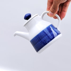 Puede incluir: Una tetera de cerámica blanca con una banda azul con un diseño de hojas en el centro. La tetera tiene un pico curvo, un asa y un botón azul en la tapa. La tetera está sujeta por una mano.