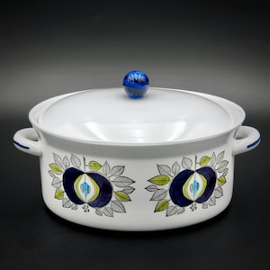 Puede incluir: Plato de cerámica blanca para hornear con tapa de pomo azul. El plato tiene un patrón floral con hojas azules, verdes y negras y fruta morada.