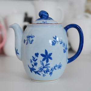 May include: A light blue ceramic teapot with a matching lid and handle. The teapot is decorated with blue floral patterns. The handle and the knob on the lid are a darker shade of blue. The teapot has a capacity of approximately 4 cups.