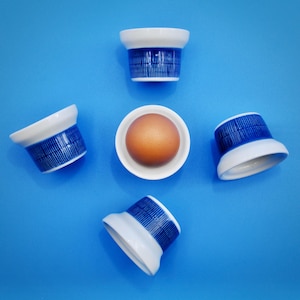 Puede incluir: Cinco hueveras de cerámica azul y blanca con un solo huevo marrón en una de las hueveras. Las hueveras tienen un patrón de rayas azules y blancas.