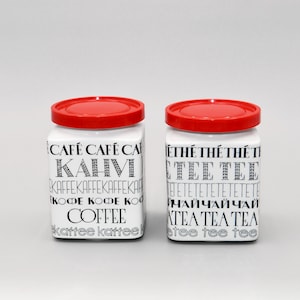 Puede incluir: Dos botes de cerámica blanca con tapas rojas. Los botes tienen texto negro en ellos, incluyendo las palabras "Coffee", "Kaffee", "Kahvi", "Tee", "Chai" y "Thé".