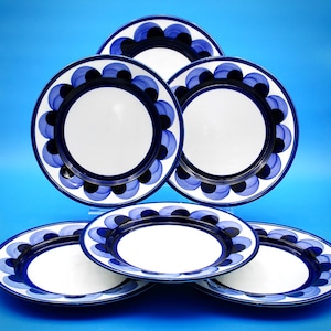 May include: A set of five round ceramic plates featuring a white center and a wide border decorated with a repeating pattern of overlapping dark blue and black circular shapes. The plates are arranged in a stacked formation against a blue background.