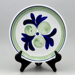 Puede incluir: Un plato de cerámica blanco con un diseño floral verde y azul. El plato tiene un borde verde y un patrón floral azul en el centro. El plato está sobre un soporte negro.