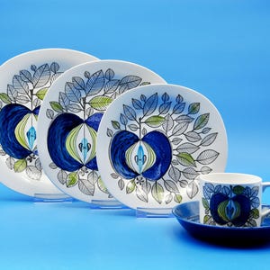 May include: A set of four white ceramic plates with a blue and green apple design. The plates are arranged in a pyramid shape with a matching teacup and saucer in front.