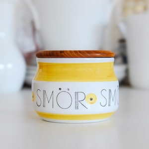 Op de afbeelding: Een witte keramische pot met gele strepen en een houten deksel. De pot heeft de tekst "SMÖR" in zwarte letters. De pot is rond en wordt waarschijnlijk gebruikt voor het bewaren van boter of andere voedingsmiddelen.