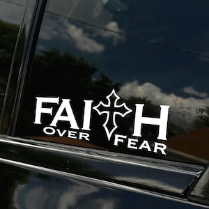 May include: White vinyl decal with the words "FAITH OVER FEAR" and a cross in the middle. The decal is on a black car window.