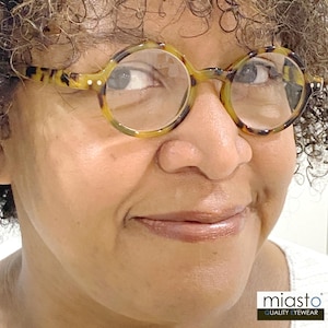May include: A close-up of a person wearing tortoise shell round glasses with a brown and black pattern. The glasses have clear lenses and a small white dot on each side of the frame. The person is smiling and looking to the right. The image includes the text "miasto" and "QUALITY EYEWEAR".