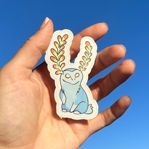 May include: A sticker featuring a whimsical blue creature with yellow and orange antler-like features. The sticker has a white border and is held against a clear blue sky. The creature has a smiling face and a light blue body.