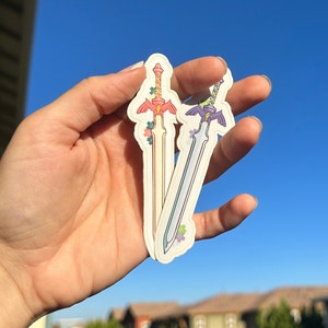 Op de afbeelding: Twee decoratieve zwaardstickers. Eén zwaard heeft een roze handvat en een crèmekleurig lemmet, versierd met roze en groene bloemaccenten. Het andere zwaard heeft een paars handvat en een zilveren lemmet, met paarse en groene bloemaccenten.