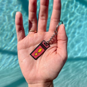 Puede incluir: Un llavero con un dije rectangular con fondo rosa y formas de diamante amarillas. El dije tiene un borde negro y está unido a una cadena y un llavero plateados. El fondo es una superficie de agua azul claro.