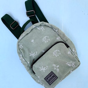 May include: A sage green corduroy backpack with a white animal pattern. The backpack has a front zipped pocket, adjustable dark green straps, and a top handle. The label reads "inti suite SUPPLY CO."