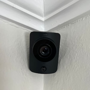 May include: A black security camera mounted in the corner of a white wall. The camera has a round lens and a small sensor on the bottom.