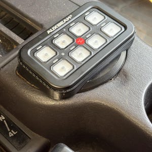 May include: A black AUXBEAM switch panel with nine buttons, including a red button labeled "+". The panel is mounted in a vehicle's dashboard.