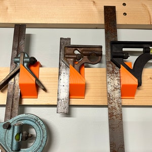 May include: A collection of vintage metal measuring tools, including squares and rulers, mounted on a wooden board with orange 3D-printed holders. The tools are in various states of wear, showcasing their age and use.