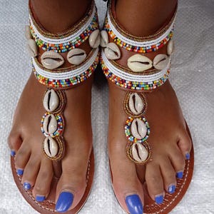 May include: Handmade leather sandals with a brown sole and straps adorned with seashells and colorful beads. The ankle straps feature white leather and gold accents. The sandals have a thong style design with seashells on the toe straps.