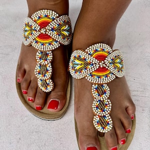 May include: Beaded sandals with a colorful design. The sandals feature a red, yellow, blue, and white beaded pattern on the straps and toe post. The footbed is a light brown color.