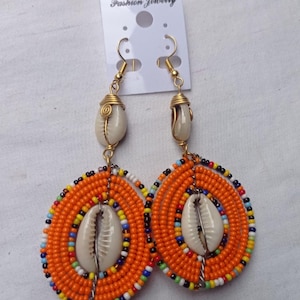 May include: A pair of handmade earrings featuring orange beaded teardrop shapes with a white seashell in the center. The earrings have gold-colored hooks and are attached to a white card that reads "Fashion Jewelry".