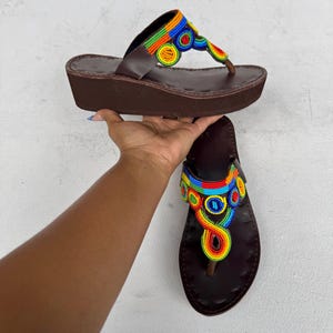 May include: Brown leather sandals with colorful beaded accents. The sandals have a wedge heel and a thong-style design. The beaded design features a rainbow of colors, including red, orange, yellow, green, blue, and purple.