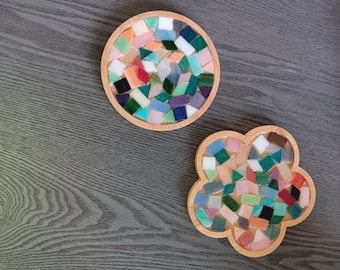 May include: Two round coasters made of wood with a mosaic design. The coasters are decorated with small, colorful glass tiles in shades of green, blue, pink, and white. The coasters are perfect for adding a touch of color and style to any home.
