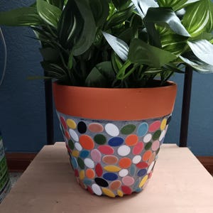 May include: A decorative flower pot with a mosaic design. The pot is filled with green leafy plants. The mosaic is made of colorful, oval-shaped tiles in shades of red, blue, yellow, and white. The rim of the pot is orange.