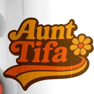 Op de afbeelding: Witte keramische mok met een retro-ontwerp met de tekst "Aunt Tifa" in oranje en bruine letters, met een bloemenafbeelding. De mok heeft een blauw handvat en rand.
