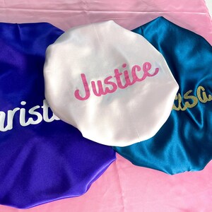 K&ouml;nnte beinhalten: Drei Hauben aus Satin in K&ouml;nigsblau, Wei&szlig; und T&uuml;rkis. Die blaue Haube hat einen wei&szlig;en Schriftzug, die wei&szlig;e Haube zeigt den pinkfarbenen Glitzertext Justice und die t&uuml;rkisfarbene Haube tr&auml;gt einen goldenen Glitzertext. Glatte, gl&auml;nzende Oberfl&auml;che.