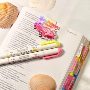 May include: Open Bible with colorful tabs and two highlighters. A pink car sticker with the text "God's Passenger Princess" is on the page. Seashells are in the background.